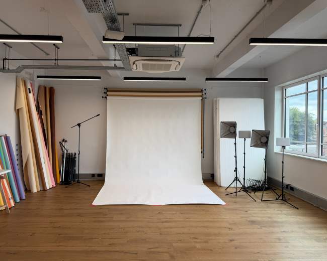The image shows a spacious photography studio with a white backdrop, lighting equipment, and colorful rolls of photographic paper against the wall.