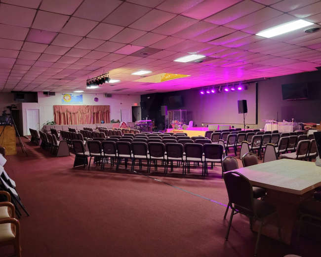 A spacious auditorium features rows of chairs facing a stage with purple lighting, flanked by sound equipment and a small band setup.