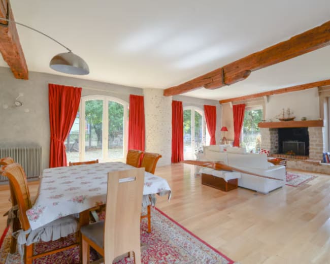 A spacious living room features a dining area with a table, a white sofa near a fireplace, and large windows dressed with red curtains.