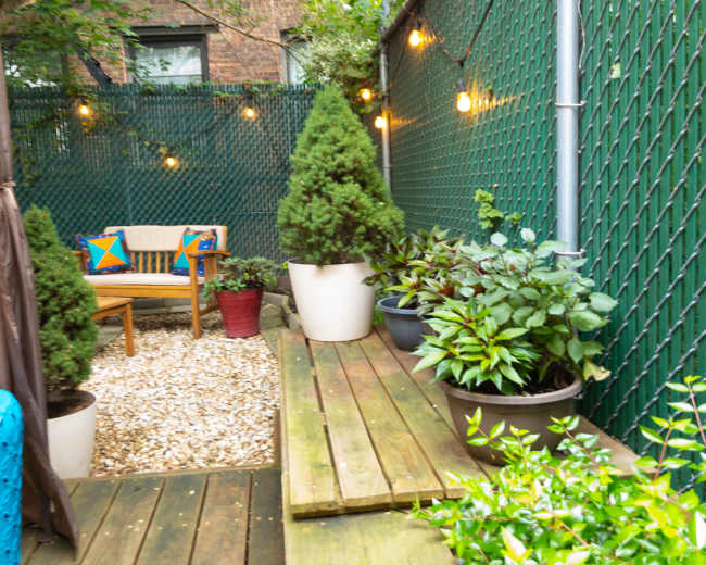 A small outdoor garden features a wooden deck with potted plants, a seating area, and string lights along a green fence.