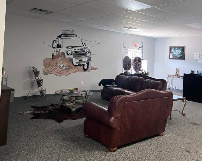 The image shows a seating area with a brown leather couch and a coffee table, featuring a mural of a vehicle on the wall and decorative items scattered throughout the room.
