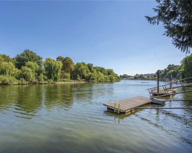 The image shows a calm river scene with a wooden dock extending into the water, surrounded by lush green trees under a clear blue sky.