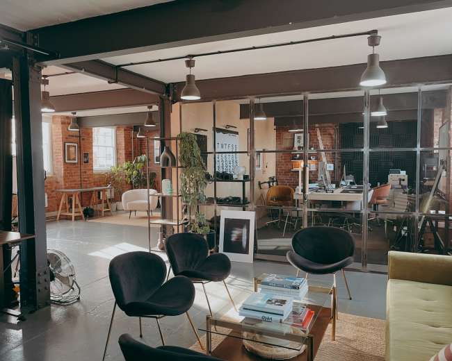 The image shows a modern office space featuring a lounge area with black chairs, a green couch, and large glass partitions separating workstations and meeting areas.
