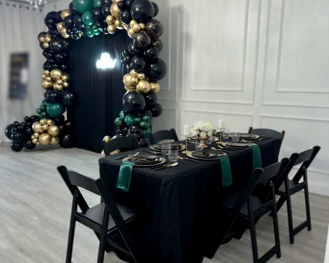 A black table with elegant dinnerware is set up in front of an arch made of balloons in black, gold, and green.