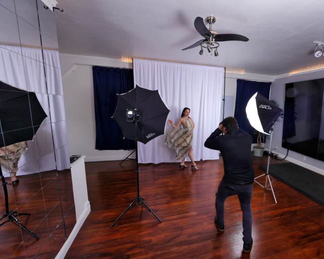A photographer is capturing images of a model posing in front of a backdrop, with studio lighting equipment set up around them.