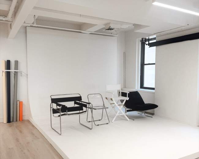 The image shows a minimalist studio space with a white backdrop and three chairs on a light wooden floor.