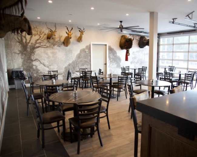 The image shows a dining area with multiple round tables and chairs arranged, complemented by a mural and mounted animal heads on the walls.