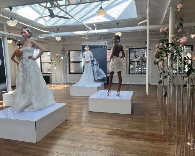 The image shows a spacious gallery featuring three mannequins dressed in elaborate bridal gowns, displayed on white pedestals surrounded by floral arrangements.