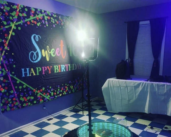 The image shows a birthday celebration setup in a room featuring a colorful backdrop, a light stand, and a decorated table with a checkered floor.