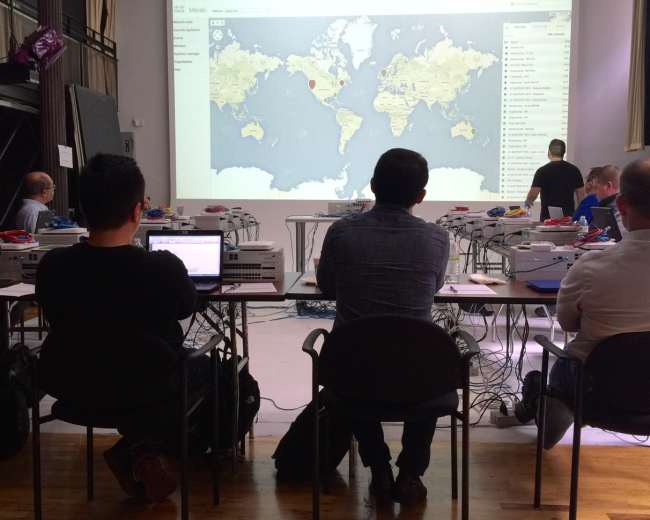 A group of people sits at a long table facing a projected world map on a screen in a classroom setting.