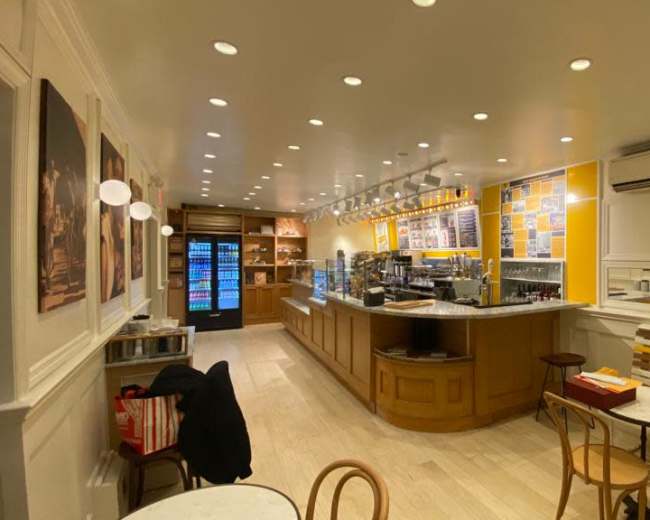 The image shows a modern café interior with a long counter, a display case, and seating arrangements, featuring bright lighting and minimalistic decor.