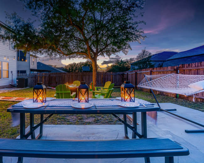 A backyard scene features a picnic table with lanterns, green chairs, a hammock, and a tree, set against a sunset sky.