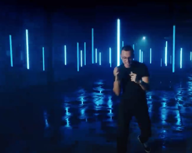 A person stands in a dimly lit space illuminated by vertical blue neon lights, with reflections on a wet floor.