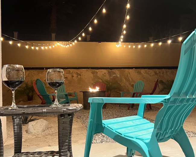 Two glasses of red wine sit on a wicker table next to a turquoise chair, with string lights illuminating a backyard featuring fire and colorful chairs in the background.