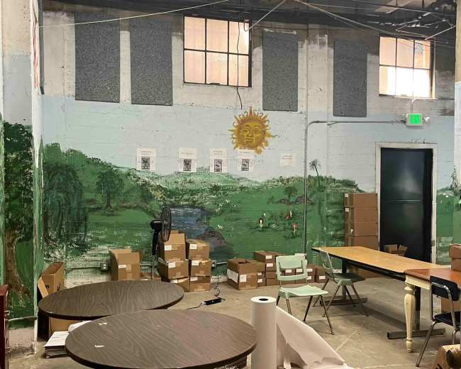 The image shows a partially furnished room with a mural of a landscape on the wall, surrounded by cardboard boxes and tables.