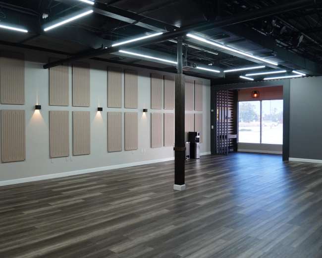 The image depicts an empty room with wooden flooring and sound-absorbing wall panels, illuminated by overhead lights.