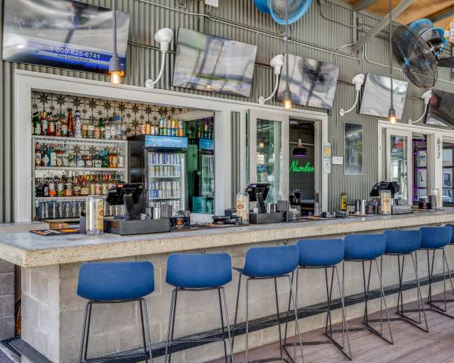 A bar with a long counter, several high stools, and shelves stocked with various bottles of liquor is set against a backdrop of large flat-screen TVs and ceiling fans.