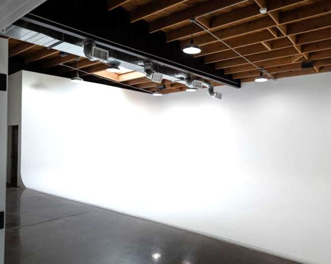 The image shows a spacious studio with a curved white backdrop and wooden ceiling beams, illuminated by overhead spotlights.