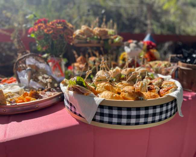 A wooden tray filled with assorted sandwiches is displayed on a pink tablecloth, surrounded by colorful flowers and additional food items.