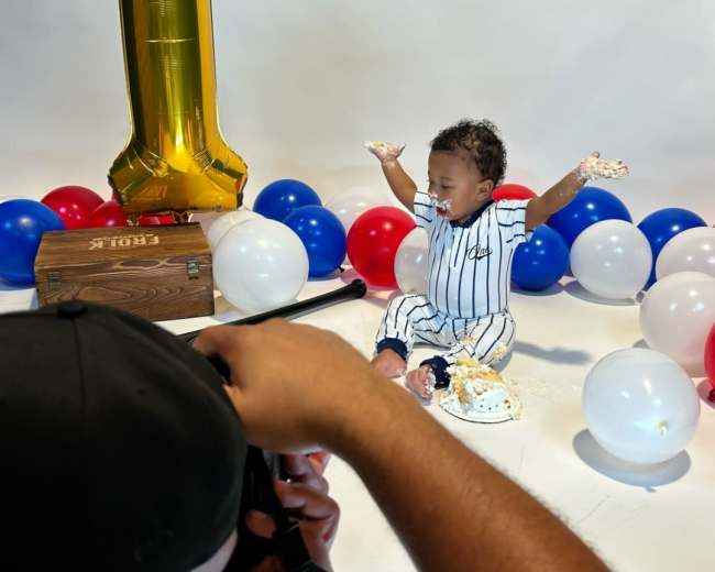 A one-year-old child sits amongst balloons and a cake, with a photographer capturing the moment.