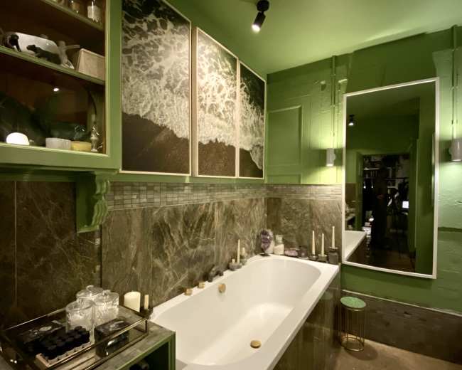 A modern bathroom features a white bathtub, green walls, and nature-themed artwork, with shelves displaying candles and toiletries.