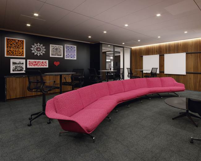 The image shows a modern office space with a large pink curved sofa, dark chairs, wooden walls, and artwork on the walls.