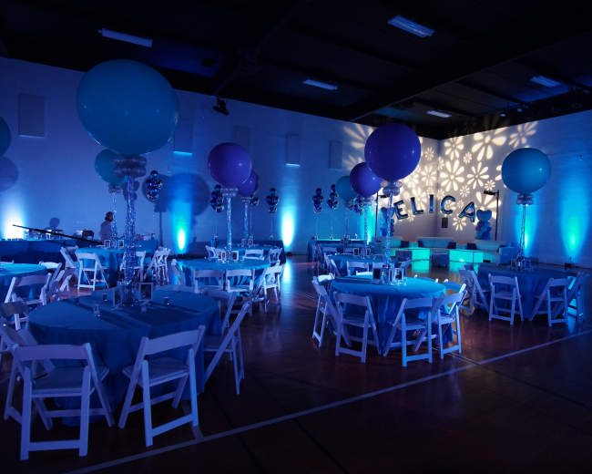 A decorated indoor venue features tables with blue tablecloths, large balloons, and illuminated walls displaying the name "FELICIA."
