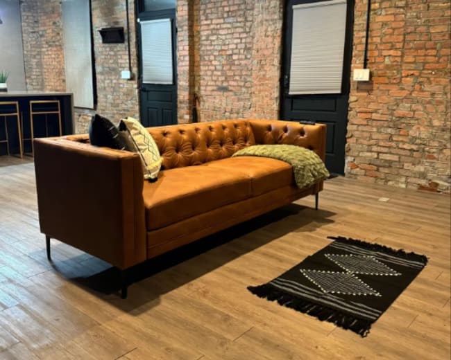 The image shows a brown tufted leather sofa positioned on a wooden floor in a room with exposed brick walls and two doors in the background.