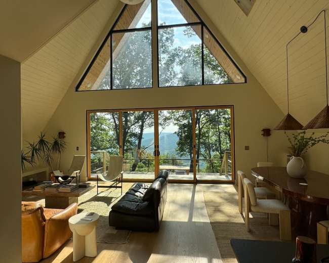 The image shows a spacious, modern living room with large windows overlooking a forested landscape.