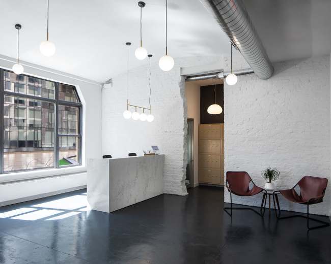The image shows a modern reception area with a white marble desk, hanging globe lights, and a seating space featuring two brown chairs and a small plant.