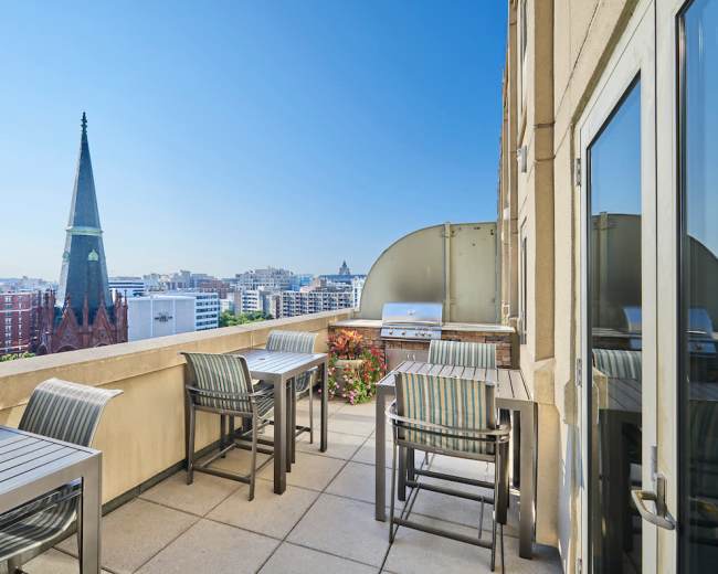 A rooftop terrace features metal tables and chairs, overlooking a skyline with a tall spire in the distance.