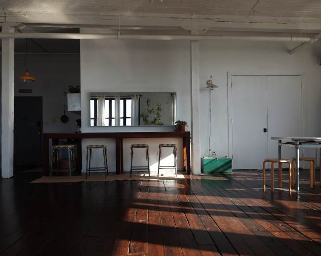 A spacious, airy interior features a long wooden bar with metal stools, a large mirror, and a dining table set against a backdrop of white walls and wooden flooring.