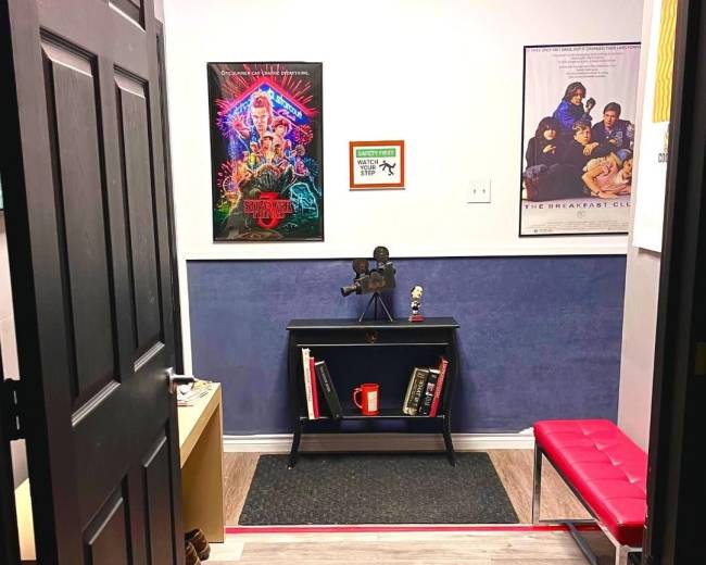 The image shows an office entrance featuring a black door, a blue wall, a small bookshelf with a red mug, and two movie posters on the walls.