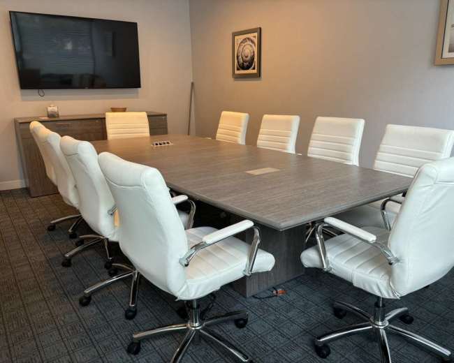 The image shows a modern conference room with a large rectangular table surrounded by eight white ergonomic chairs, a television mounted on the wall, and a side cabinet.