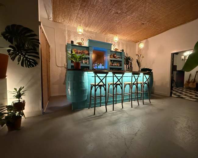 The image shows a brightly lit bar area with a turquoise bar counter, high stools, and various plants, set in a spacious room with a woven ceiling.