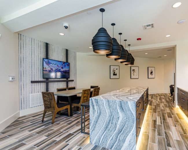 A modern kitchen area features a large island with a striped marble countertop, dark cabinetry, pendant lights, and wall-mounted television with framed artwork nearby.