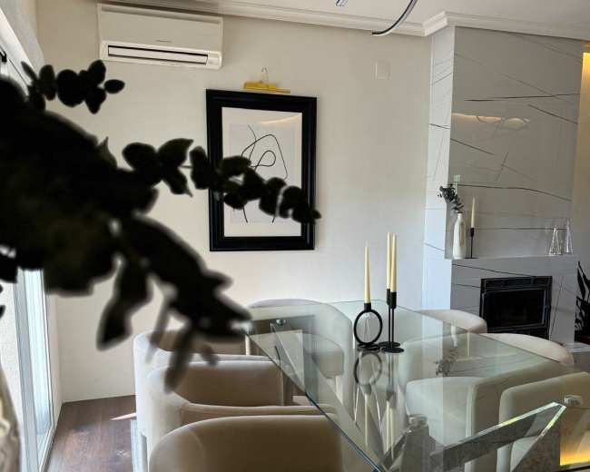 A modern dining area features a glass table surrounded by cream-colored chairs, with a black and white artwork on the wall and candles on the table.