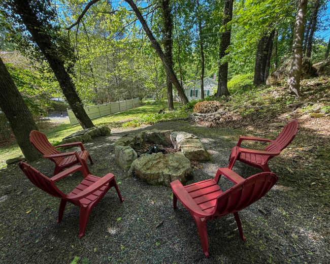 A circular arrangement of red Adirondack chairs surrounds a stone fire pit in a wooded area.