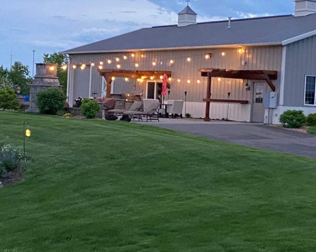 A modern barn-style building is illuminated by string lights on a landscaped lawn, featuring outdoor seating under a covered porch.