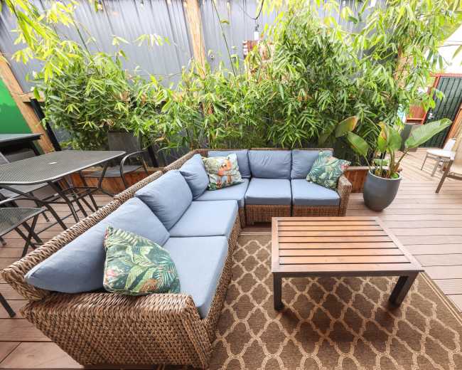 The image shows a patio area featuring a large sectional wicker couch with blue cushions, a wooden coffee table, and plants in the background.