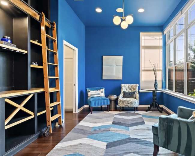 A bright room features blue walls, a geometric rug, two upholstered chairs, and a ladder against a shelving unit.