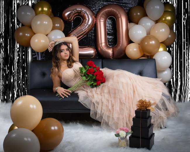 A woman in a beige gown holds a bouquet of red roses while reclining on a black couch surrounded by gold and white balloons and large "20" balloons in the background.