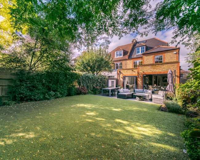 The image shows a well-maintained backyard with artificial grass, surrounded by lush greenery and featuring a two-story brick house.