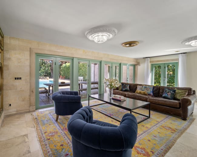 The image shows a stylish living room with a leather sofa, two blue armchairs, a glass coffee table, and large windows facing a pool area.