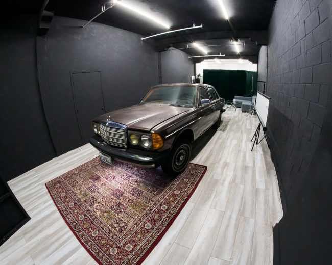A vintage car is parked on a patterned rug inside a dimly lit room with black walls and wood-like flooring.