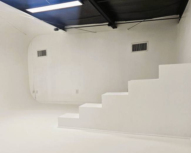 The image shows a minimalistic white room featuring a curved wall and a staircase leading to a small elevated platform.