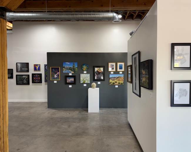 The image shows an art gallery with framed artworks displayed on a dark gray wall and additional pieces on a light-colored wall, while a pedestal in the center holds a sculpture or bust.