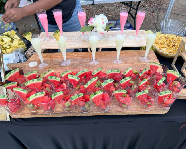 A wooden serving board featuring several glasses filled with watermelon slices, alongside colorful, decorative drinks on a table.