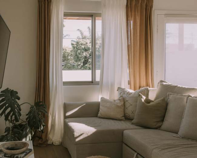 The image shows a cozy living room with a sectional sofa, a small woven pouf, and large windows covered with sheer and curtain panels.