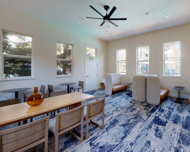 The image shows a modern common area featuring a dining table, several chairs, and lounge seating with large windows allowing natural light.